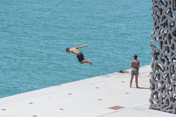 Une jeune garçon plonge du quai du MuCEM à Marseille devant le regard de ses copains.