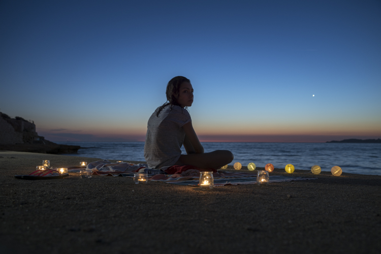 Une jeune fille assisse en tailleur sur une serviette faisant face à la mer est entourée de bougies et de guirlande lumineuse comme pour une cérémonie dincantation à une divinité ou un esprit.