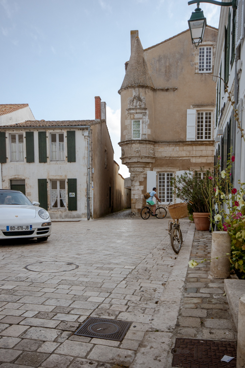 Une rue d'Ars-en-R� en fin d'apr�s-midi o� se croise v�hicule de luxe et v�lo devant une ancienne b�tisse.