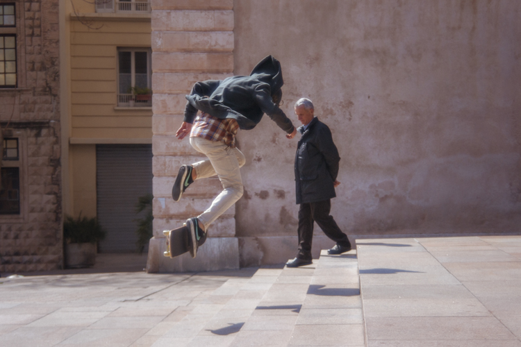Un homme descend les trois marches dun escalier tranquillement les mains dans le dos pendant quun adolescent descend ce même escalier en effectuant une figure avec son skateboard.